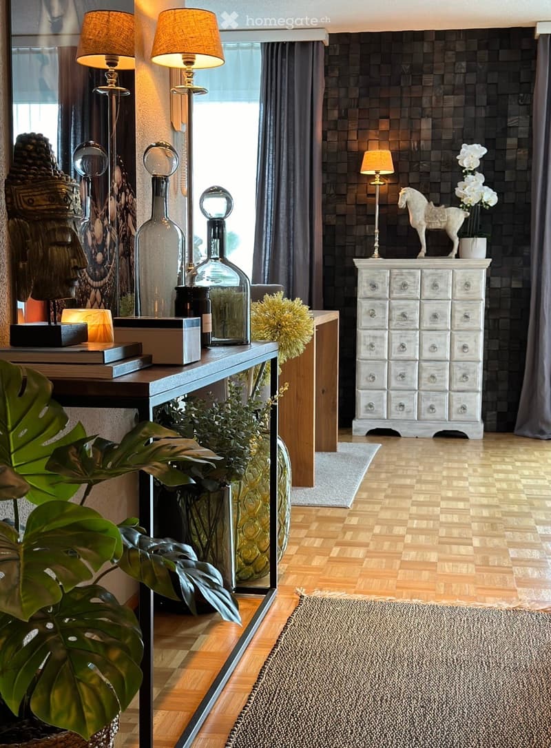 Modern hallway with wooden flooring, large mirror, decorative table, lamps, plants, and a grey cabinet.