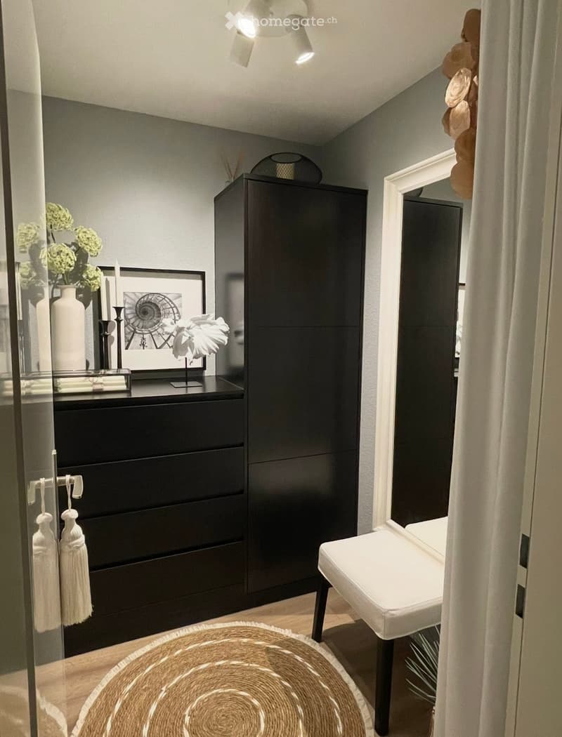 Modern, furnished room with a large wardrobe, a round woven rug, a white stool, a decorative table with a vase and picture frame, and a mirror on the wall.