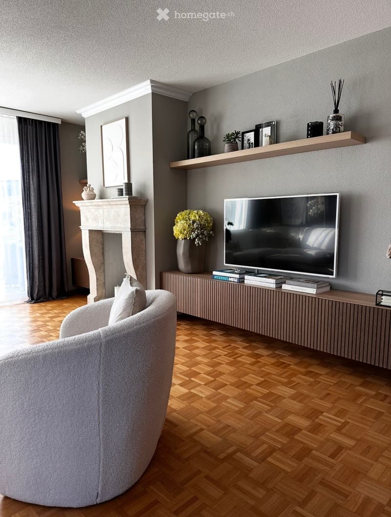 Modern living room with a round sofa, television on a wooden console table, decorative vases, books, shelves, and a fireplace.