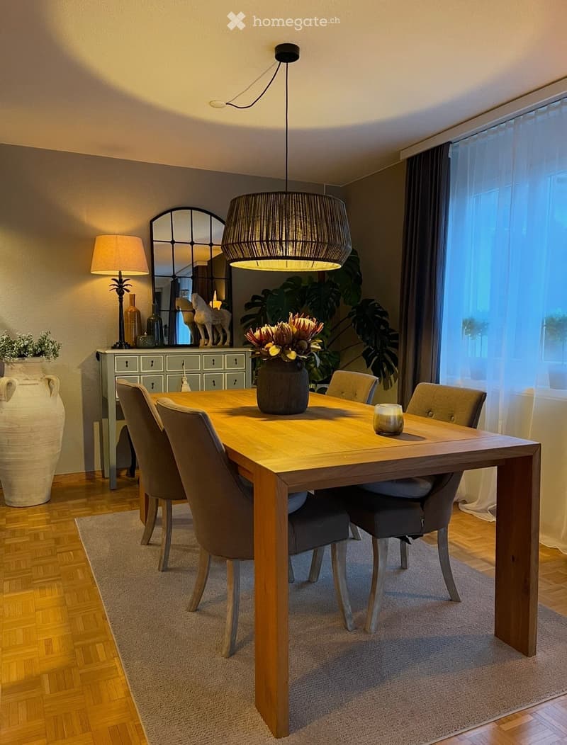 Dining room with a wooden dining table, six chairs, a large vase with flowers, a hanging lamp, a side lamp, a mirror, and a plant in the corner.