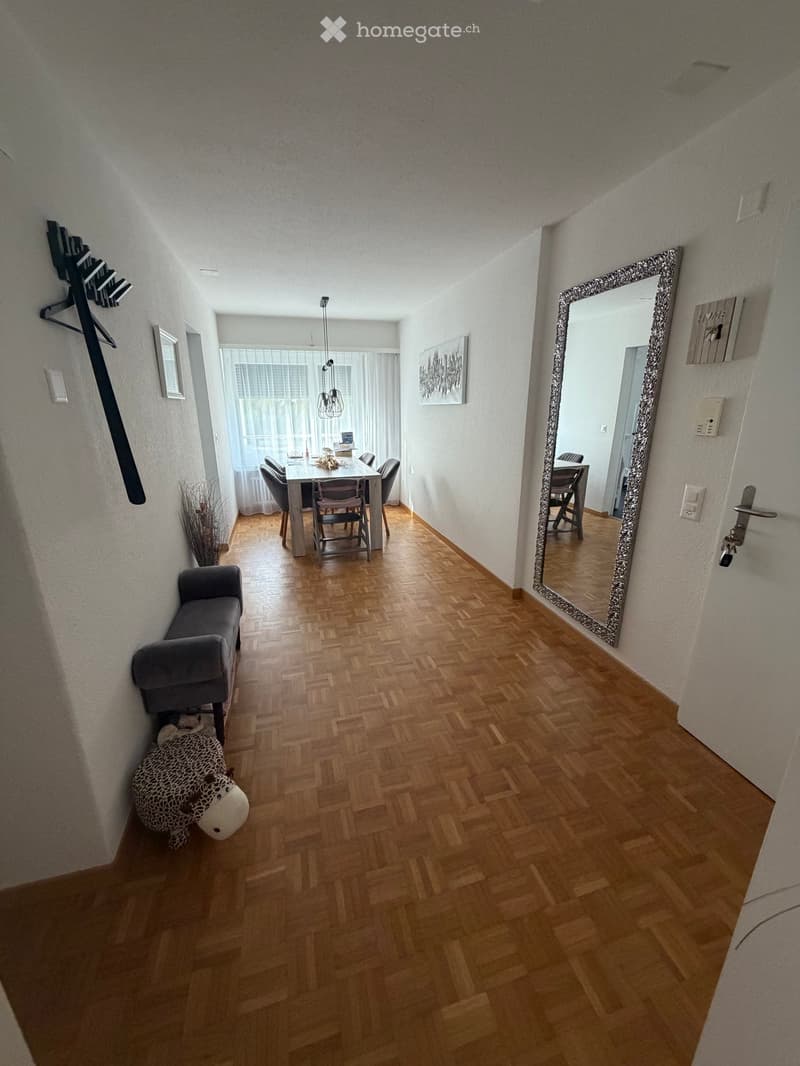 Hallway with wooden flooring, a grey bench, a large mirror, and a small decorative item on the floor. The hallway leads to a dining area with a table and chairs.