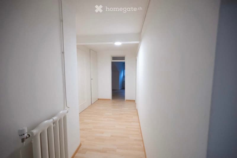 A clean, well-lit hallway with light wooden flooring, white walls, and a white radiator on the left side. The hallway leads to an open doorway at the end.