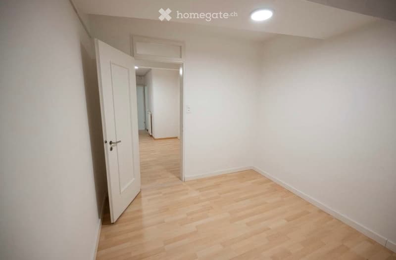 Empty hallway with light wooden flooring, white walls, and a single open door leading to another room.