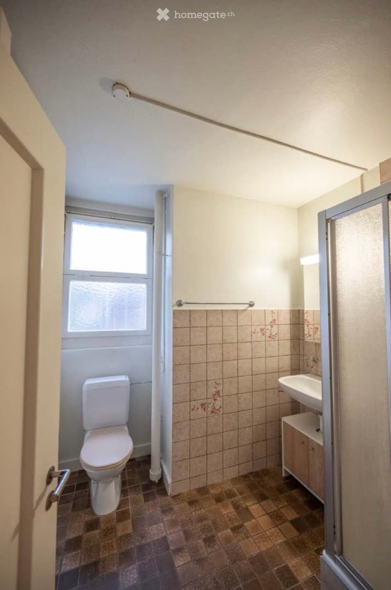 Bathroom with a toilet, sink, and cabinet. The walls are tiled with a floral pattern, and there is a window providing natural light.