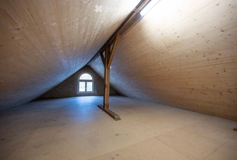 An empty attic space with wooden walls, exposed wooden beams, and a skylight window, featuring a smooth concrete floor.