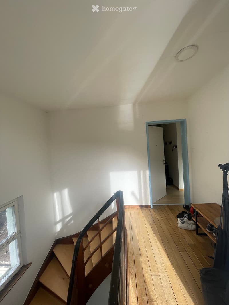 A narrow hallway with wooden flooring, white walls, and a staircase leading upwards. A small shelf holds shoes and a bag, and a window on the left allows natural light to enter.