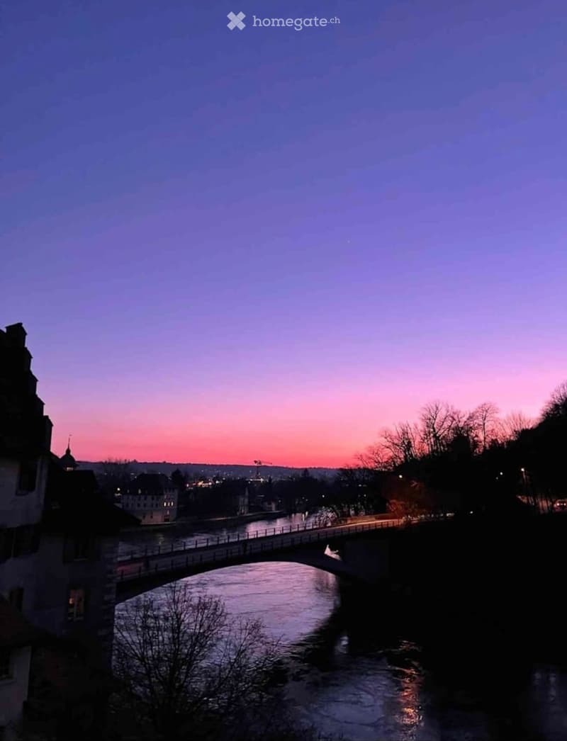 A serene riverside scene at sunset, featuring a bridge spanning the water, with silhouettes of trees and buildings on either side.