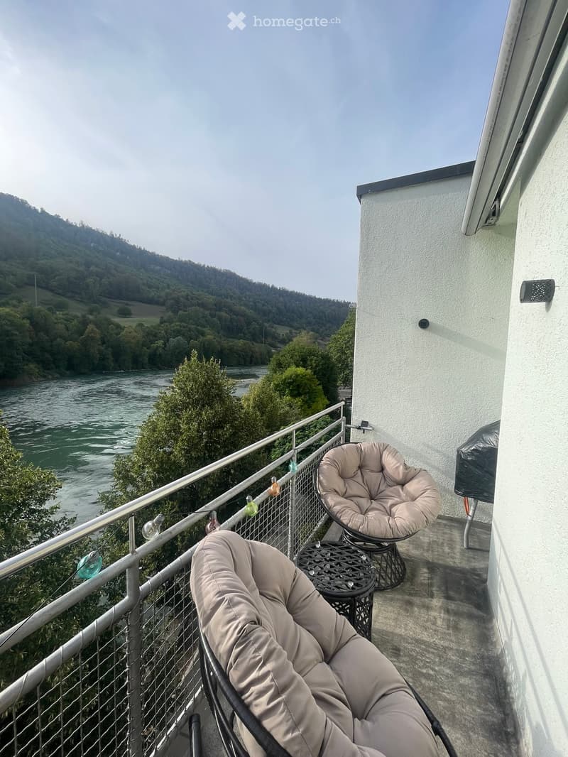 Balcony with two beige chairs and a small black table, overlooking a river and green landscape.