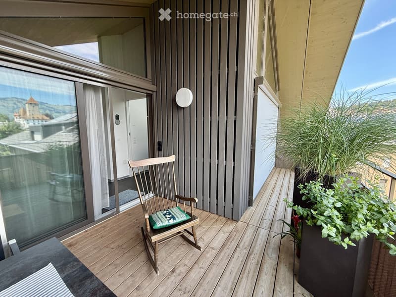 Balcony with wooden decking, rocking chair with cushion, potted plants, and scenic city view.