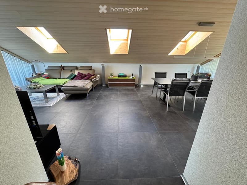 Modern living and dining area with grey sofa, coffee table, dining table, chairs, and a large screen. The space features skylights and a sleek design.