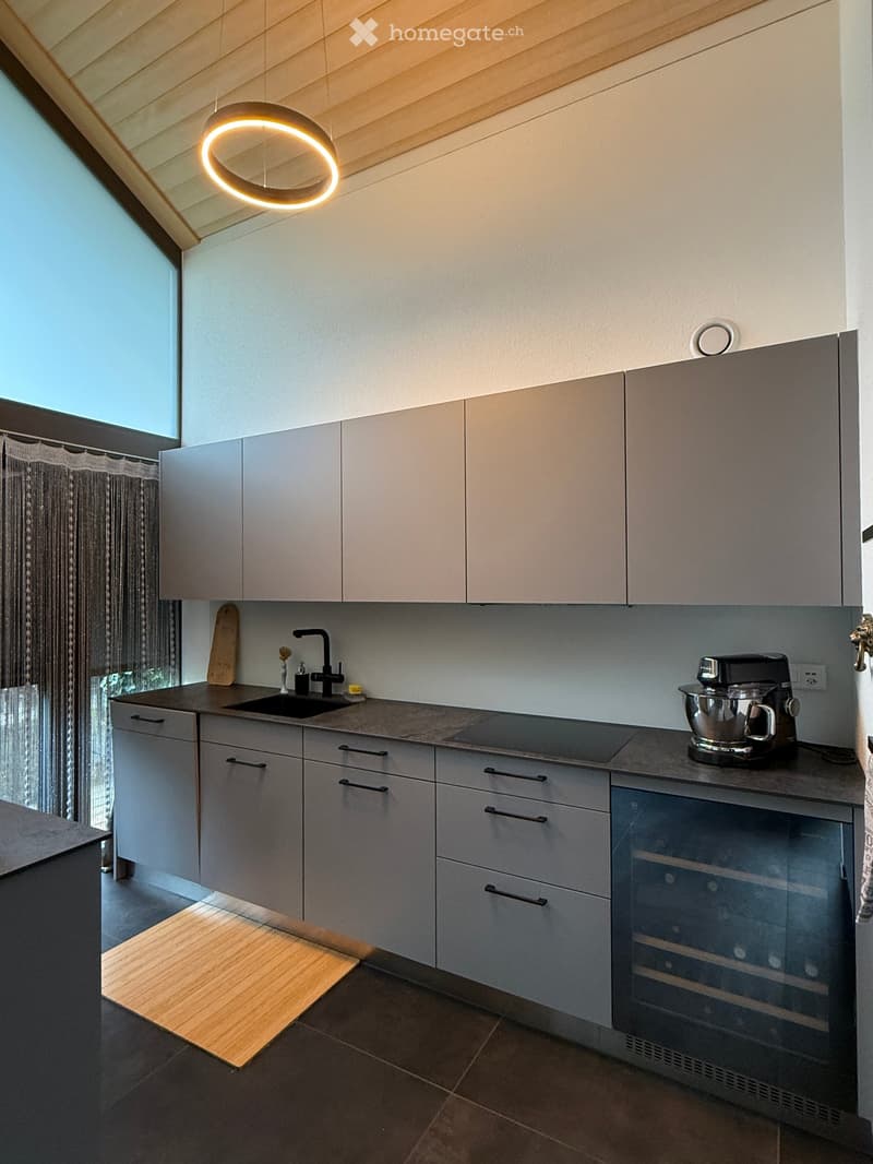 Modern kitchen with grey cabinets, a sink, a mixer, and a wine cooler. Features a large window with a curtain and a circular light fixture on the ceiling.