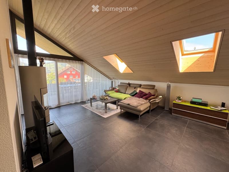 Modern living room with a slanted wooden ceiling, large windows, and skylights. Features a beige sectional sofa with colorful cushions, a coffee table, a side table with a potted plant, and a television on a stand.