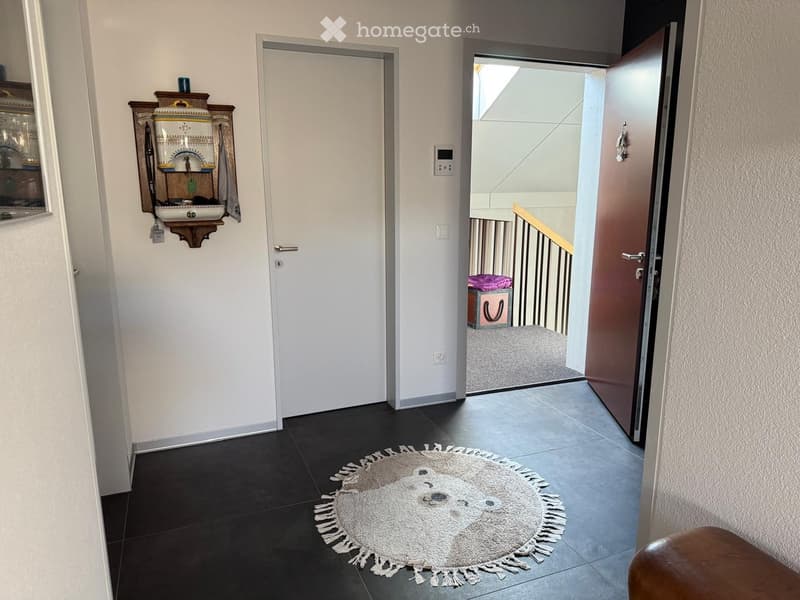 Hallway with dark tiled floor, white walls, and a round rug with a teddy bear design. There are two doors, one leading to a staircase, and a decorative wall-mounted item on the left side.