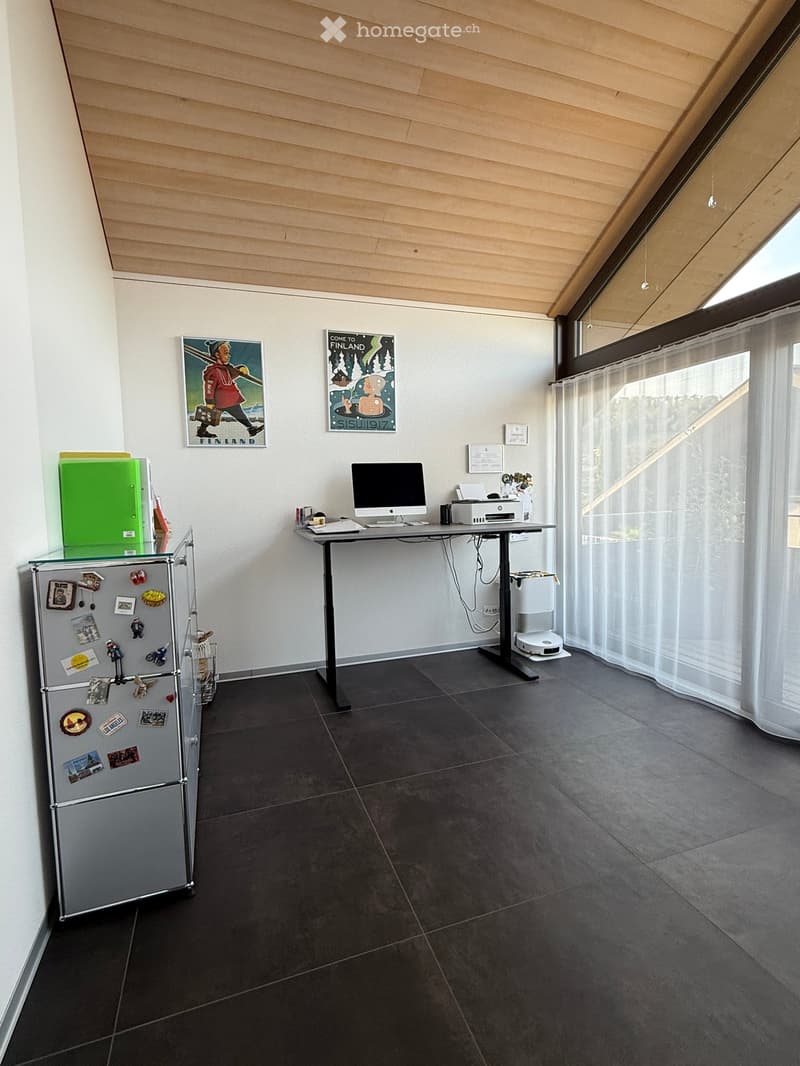 Modern, minimalist room with dark tiled floor, white walls, large window with white curtain, a small table with a computer and printer, a refrigerator with magnets, and two posters on the wall.