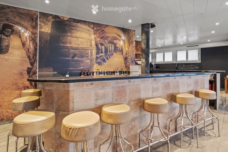 Modern bar area with a stone countertop, multiple bar stools, and a variety of wine bottles displayed on the counter. The backdrop features a large mural of a wine cellar with barrels.