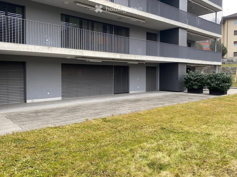 Modern multi-story building with grey facade, large garage doors, balconies, and small garden area with shrubs.
