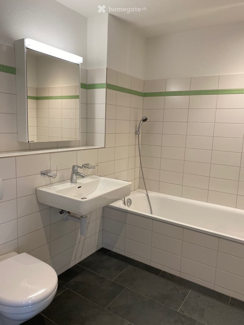 Modern bathroom with white tiles, a bathtub, a sink, a toilet, and a mirror with a built-in light. Green accent tiles line the walls.