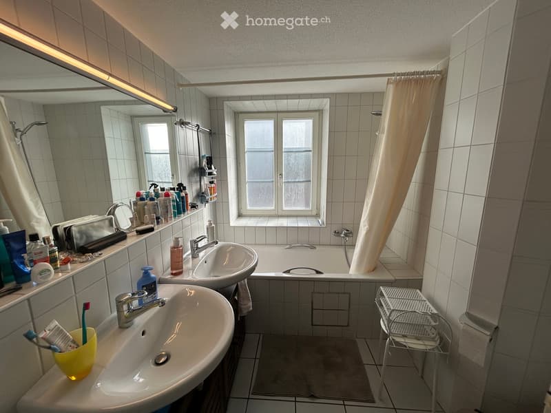 Bathroom with two sinks, a bathtub, a large mirror, and various toiletries on the counter. The room features tiled walls and a window with frosted glass panes.