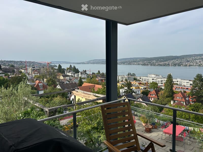 Balcony with wooden chair, red table, potted plants, and a scenic view of a city and lake.