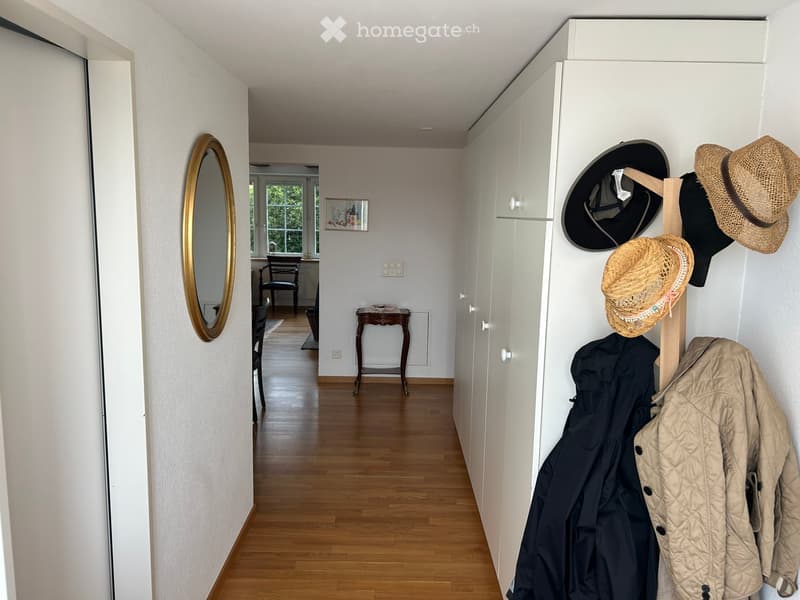Hallway with wooden flooring, white walls, a round mirror with a gold frame, a coat rack with hats and jackets, and a small table with a chair in the background.