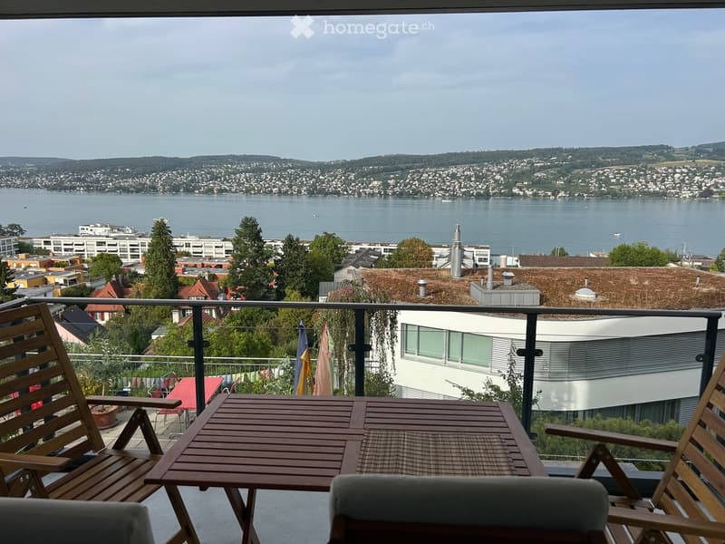 Balcony with wooden table and chairs, overlooking a scenic lake and cityscape.