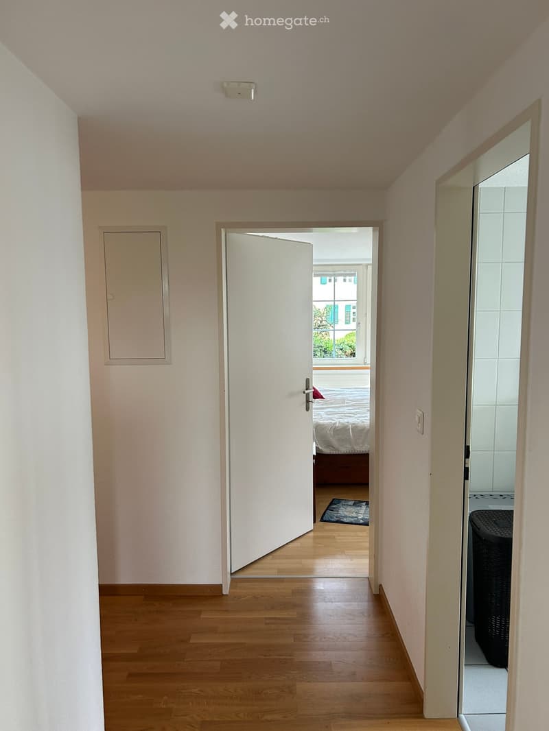 A narrow hallway with light wooden flooring and cream-colored walls, leading to a bedroom and a bathroom. The bedroom has a bed, a rug, and a window with a view of the outside. The bathroom has a visible toilet and a mirror.