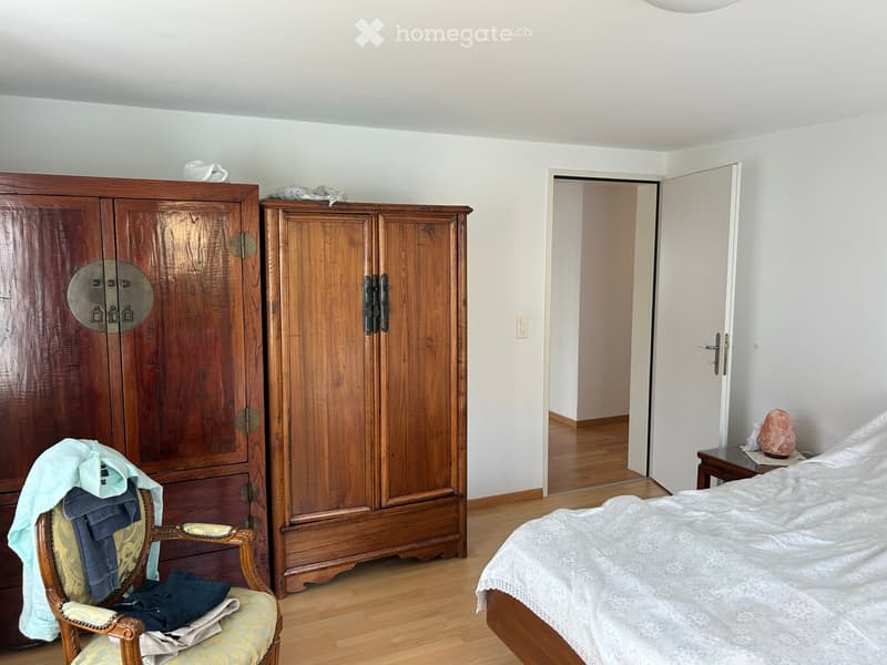 A well-lit bedroom with wooden flooring, a large bed with white bedding, a wooden wardrobe, a patterned armchair with folded clothes, and a small table with a decorative item.