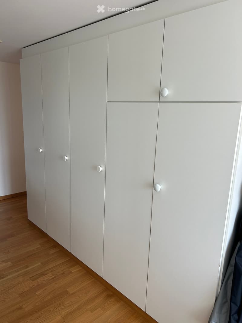 Empty room with light-colored wooden flooring and white built-in wardrobes with multiple doors and handles.