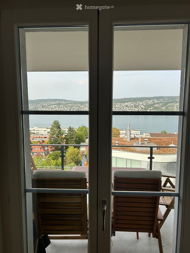 Balcony with wooden chairs and a table, overlooking a scenic view of a city and water.