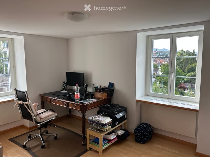 A well-lit study or home office with a wooden desk, ergonomic chair, laptop, printer, and various office supplies. Large windows offer a scenic view of greenery and buildings.