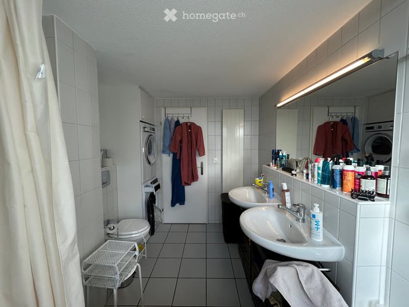 Modern bathroom with two sinks, a toilet, a washing machine, and a mirror. The space features tiled walls and floors, with personal care items and a bathrobe visible.