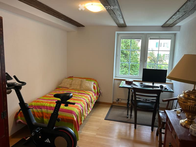 Cozy bedroom with a small bed, desk, chair, exercise bike, and a lamp near the window.