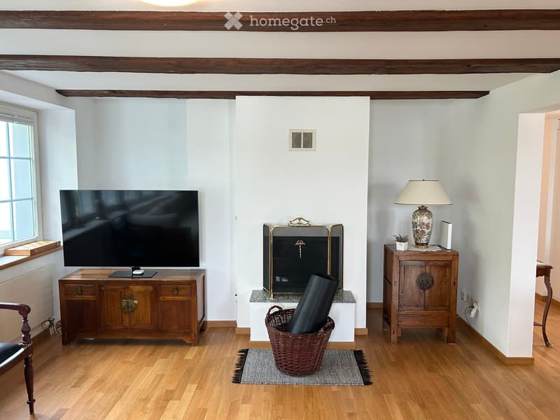 Living room with wooden floors, exposed wooden beams, fireplace, television on wooden cabinet, table lamp, and wicker basket with items.