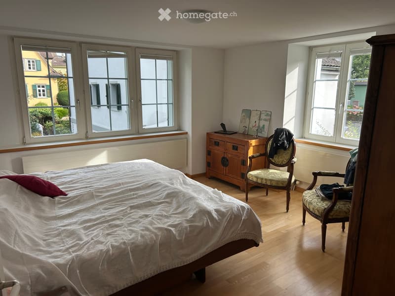 Bright bedroom with wooden flooring, large windows, a neatly made bed with white bedding and a red pillow, a wooden cabinet with drawers, two ornate chairs, and a laptop on the cabinet.