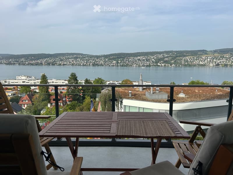 Balcony with wooden table and chairs, overlooking a scenic lake and cityscape.