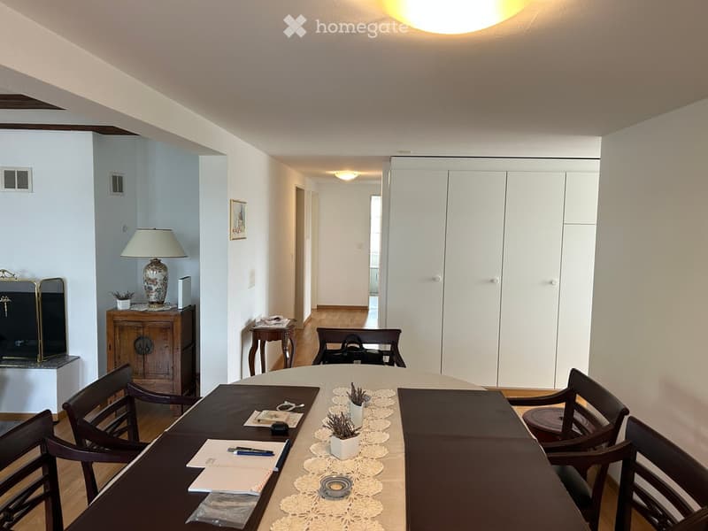A furnished room with a round wooden table surrounded by chairs, a lamp on a side table, a cabinet, and a mirror. The room has light-colored walls and wooden flooring.
