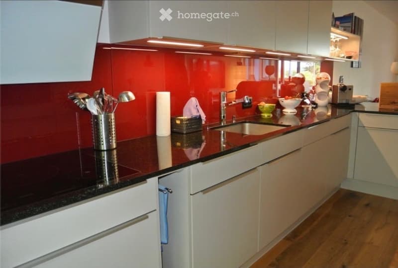 Modern kitchen with white cabinets, dark countertop, red backsplash, and stainless steel sink. Includes a knife block, paper towel holder, and small appliances on the counter.