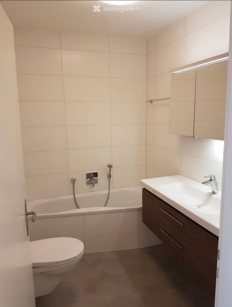 Modern bathroom with a bathtub, toilet, sink, and dark wooden cabinet. The walls are tiled in light beige, and the floor is concrete.