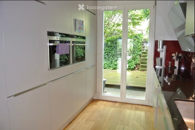 Modern kitchen with white cabinets, stainless steel oven, and a large glass door leading to a garden view.