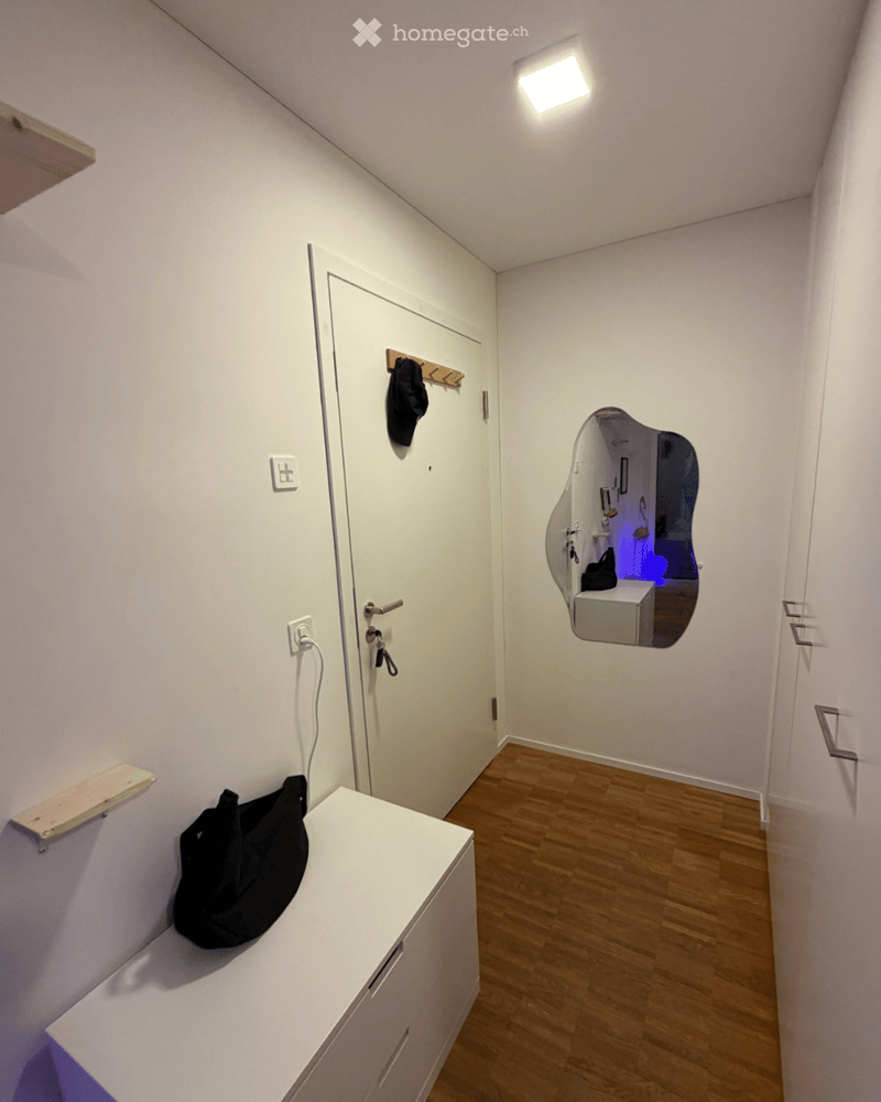 A narrow hallway with wooden flooring, white walls, and a white cabinet with a black bag on top. A door leads to another room, and a mirror is mounted on the wall.