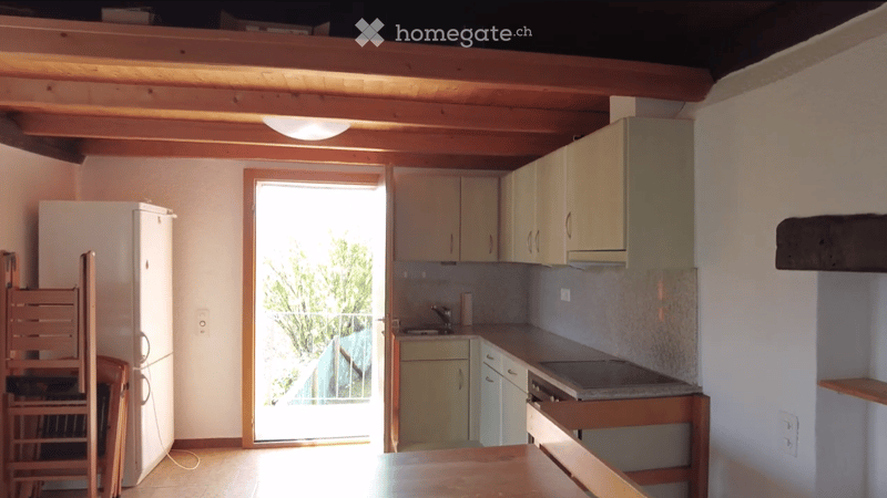 Bright kitchen with light-colored cabinets, a refrigerator, a sink, and a stove. A wooden table is visible, and there is a view of greenery through an open doorway.