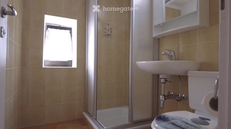 Bathroom with beige tiled walls, a white sink with a chrome faucet, a toilet, a mirror above the sink, and a glass-enclosed shower stall.