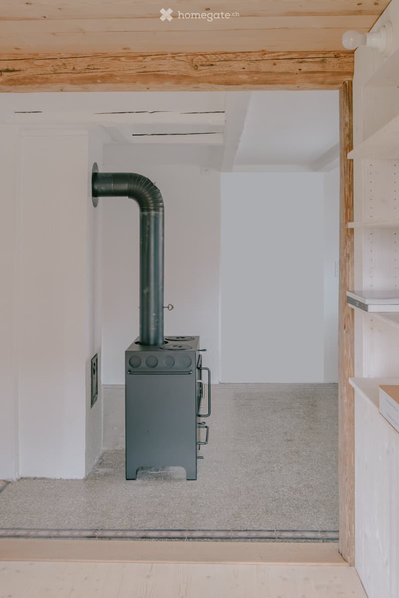 Empty room with a modern wood-burning stove, exposed wooden ceiling beams, and a mix of concrete and wooden flooring.