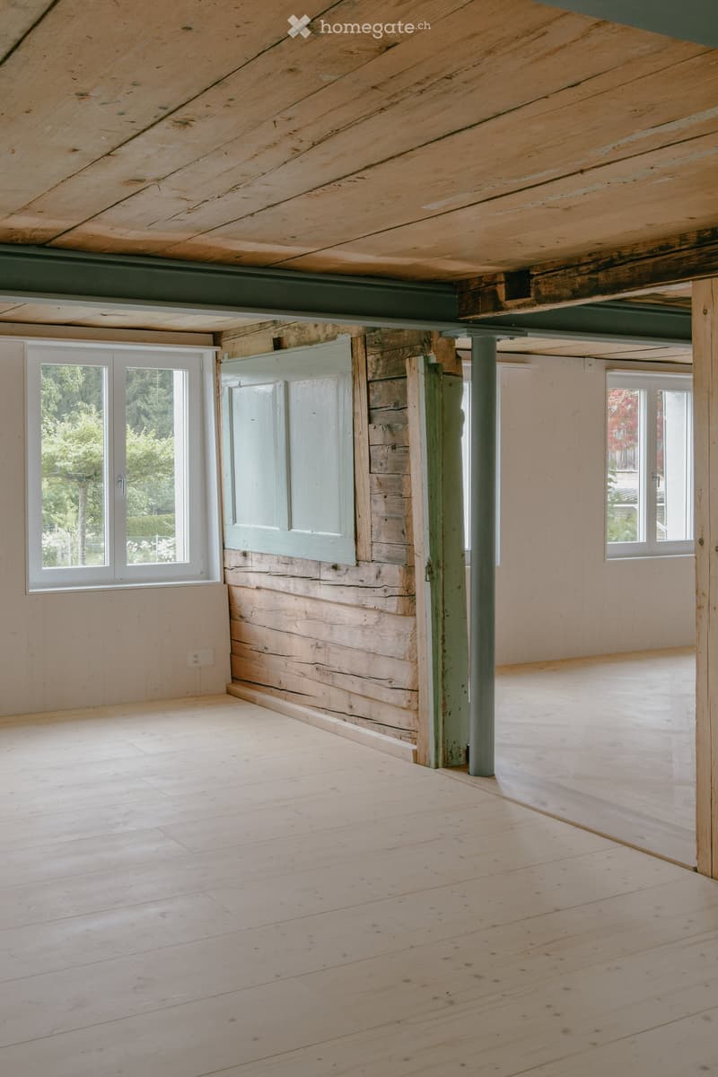 Empty room with wooden floors, exposed wooden beams, and large windows allowing natural light. The walls are painted in a light color, and there are structural columns supporting the ceiling.