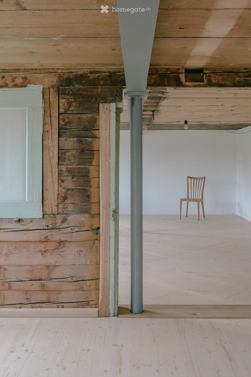 Minimalist room with exposed wooden beams, a single wooden chair, and a central column. The space features light wooden flooring and white walls.