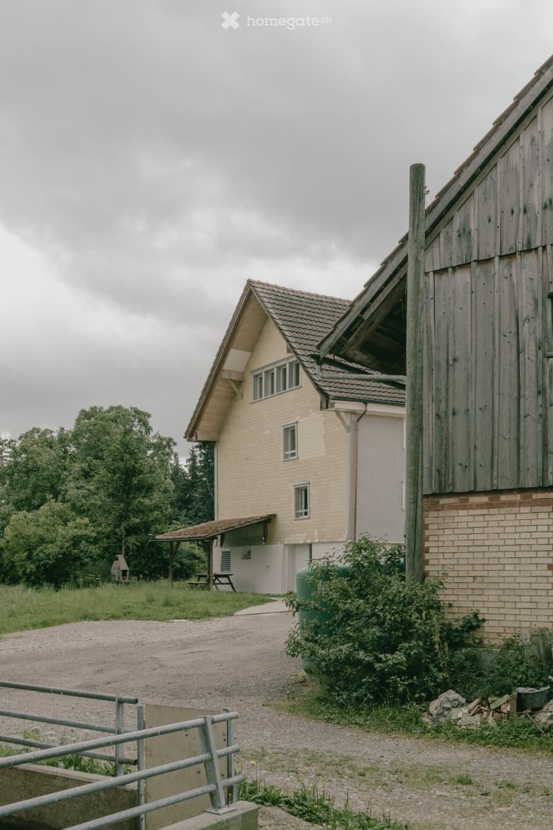 A two-story building with a beige facade and a wooden barn adjacent to it. The building has multiple windows and a covered entrance area. There is a gravel driveway leading to the building, surrounded by greenery and a metal fence.