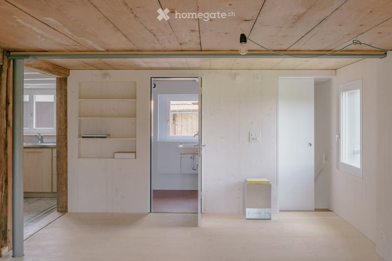 Bright, minimalist room with light wooden walls and ceiling, featuring a small sink area visible through an open doorway, shelves with books and boxes, and a small glass stool.
