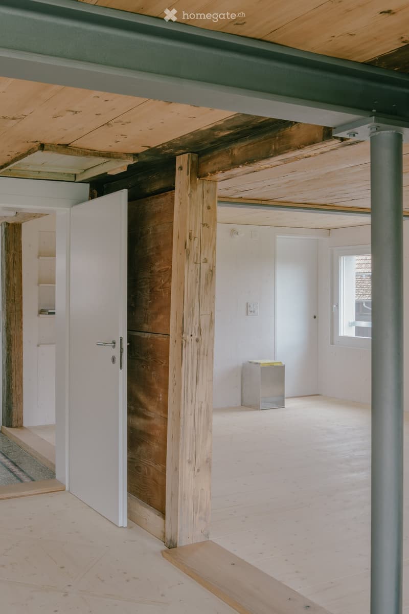An empty room with exposed wooden beams and a raised wooden floor, featuring a white door and a small window.