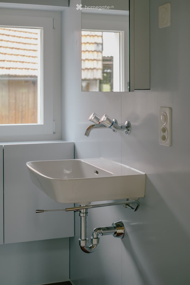 Modern bathroom with a white sink, chrome faucet, and a mirror above it. The sink is mounted on a wall with visible pipes and a towel rack below.
