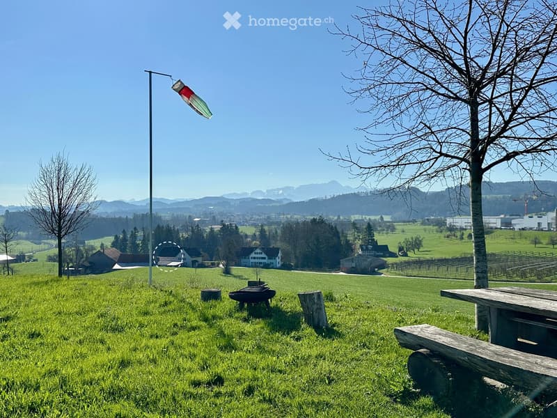 A serene outdoor area with a grassy field, picnic table, benches, and a scenic view of mountains and houses in the distance.
